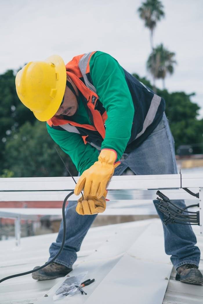 A construction worker wearing PPE installs electrical equipment on a roof.